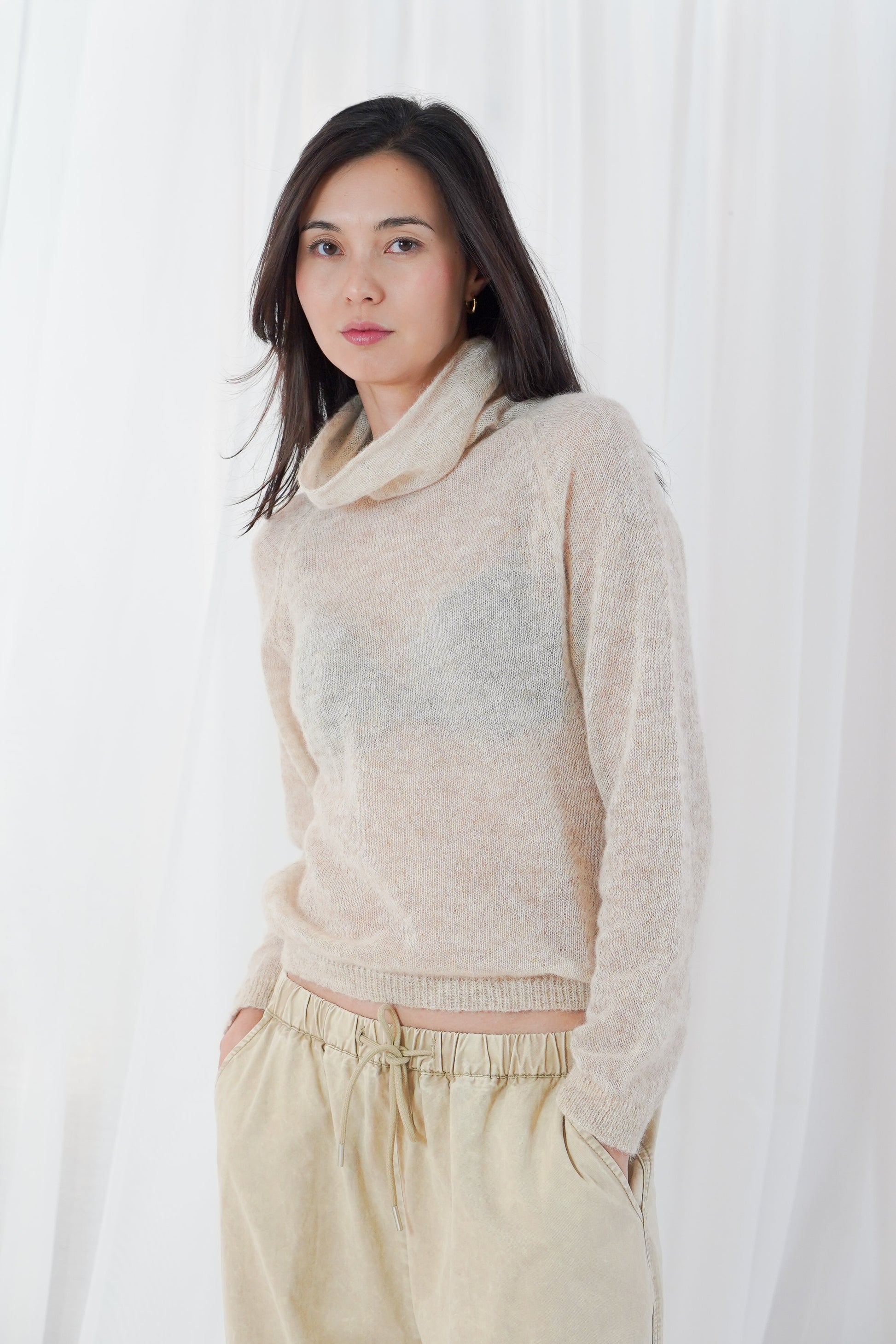A woman wearing a fuzzy, light beige cowl-neck sweater and matching beige drawstring trousers, standing against a white curtain backdrop.