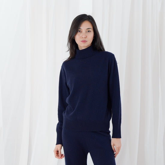A woman modeling a dark navy blue knit turtleneck sweater and matching wide-leg pants against a white draped fabric backdrop.