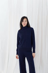 A woman modeling a dark navy blue knit turtleneck sweater and matching wide-leg pants against a white draped fabric backdrop.