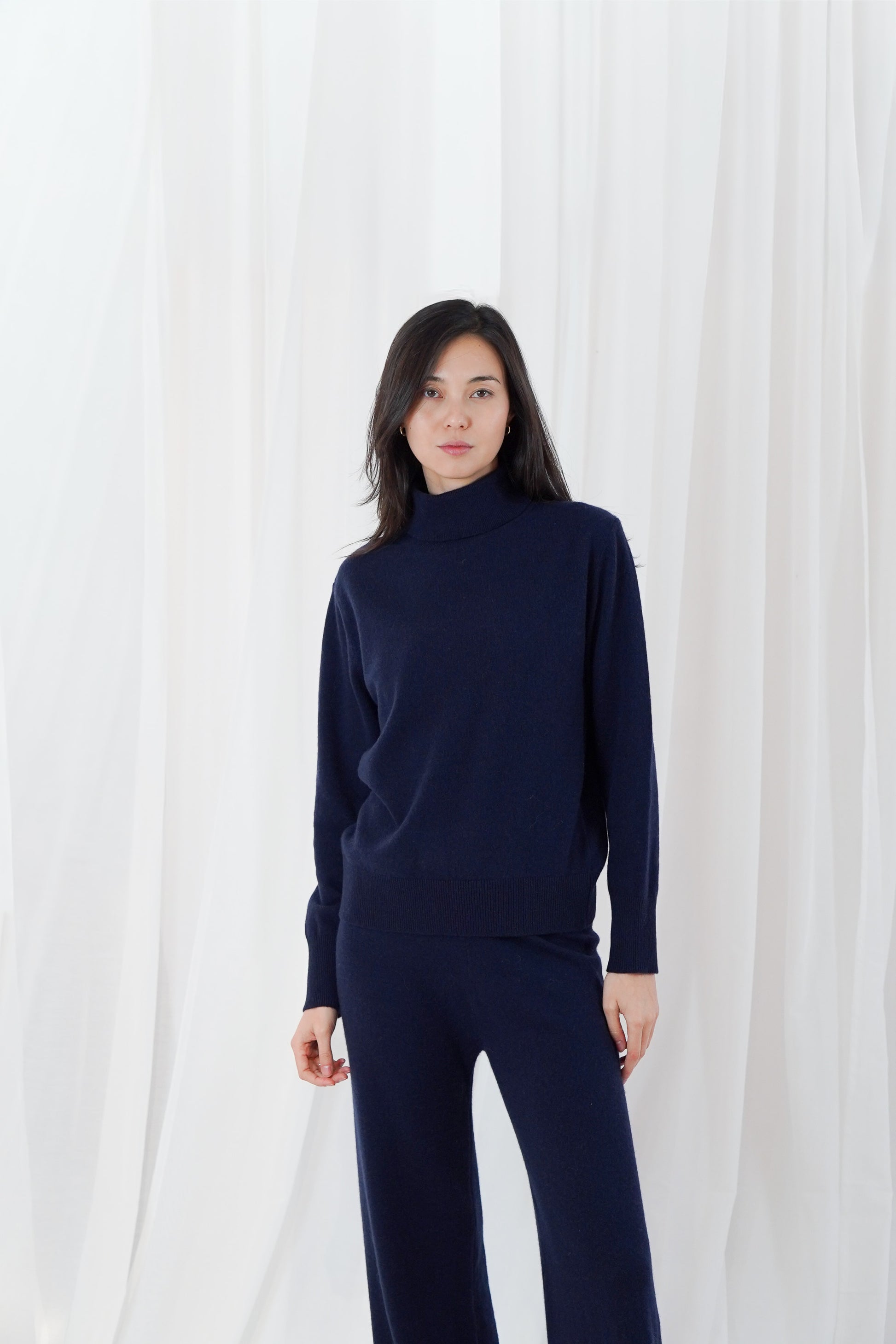 A woman modeling a dark navy blue knit turtleneck sweater and matching wide-leg pants against a white draped fabric backdrop.