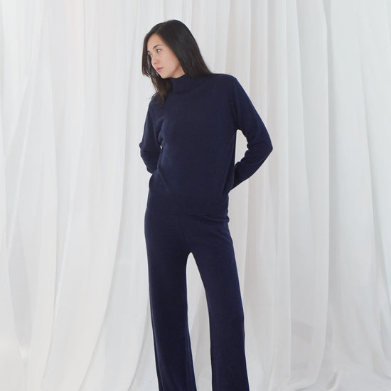 A woman modeling a navy blue mock neck jumpsuit with wide-leg pants, standing against a draped white fabric backdrop.