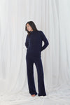 A woman modeling a navy blue mock neck jumpsuit with wide-leg pants, standing against a draped white fabric backdrop.