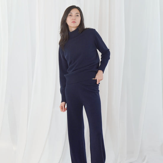 A woman modeling a matching navy blue knit loungewear set consisting of a mock-neck long-sleeve top and wide-leg pants, standing against a white draped fabric backdrop.
