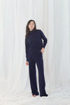 A woman modeling a matching navy blue knit loungewear set consisting of a mock-neck long-sleeve top and wide-leg pants, standing against a white draped fabric backdrop.