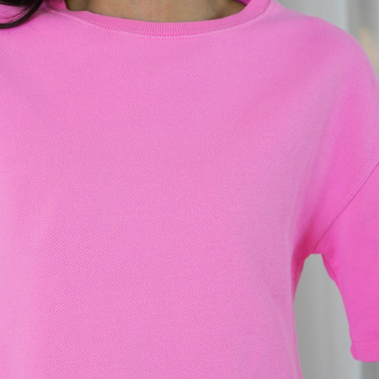 A close-up of a person wearing a bright pink short-sleeve crewneck top, showing the neck, collar, and shoulder area against a light neutral background.
