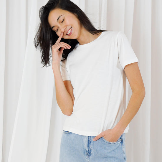 A woman wearing a plain white short-sleeve crewneck t-shirt and light blue jeans, standing in front of a white curtain background.