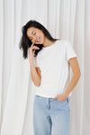 A woman wearing a plain white short-sleeve crewneck t-shirt and light blue jeans, standing in front of a white curtain background.