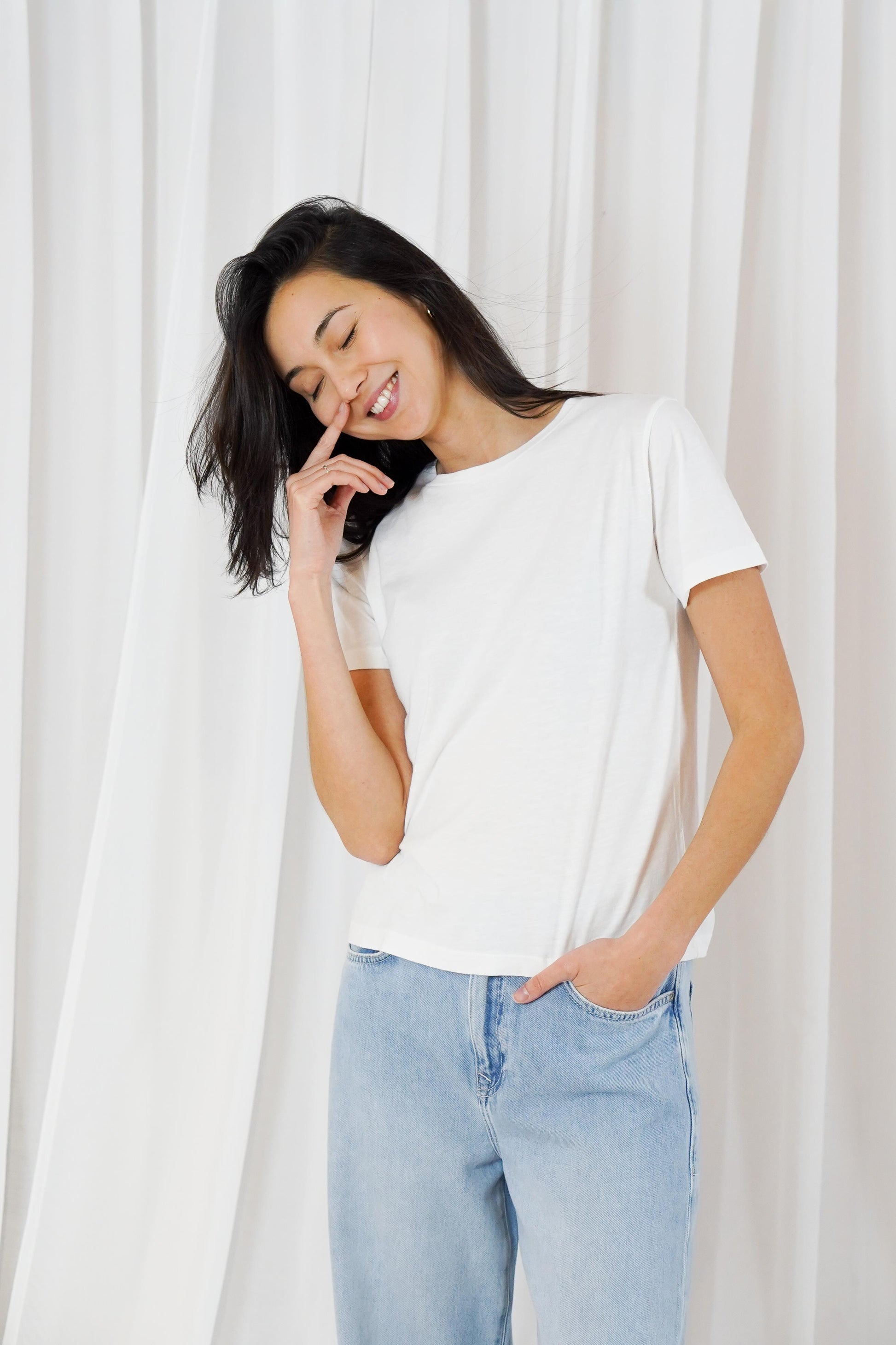 A woman wearing a plain white short-sleeve crewneck t-shirt and light blue jeans, standing in front of a white curtain background.