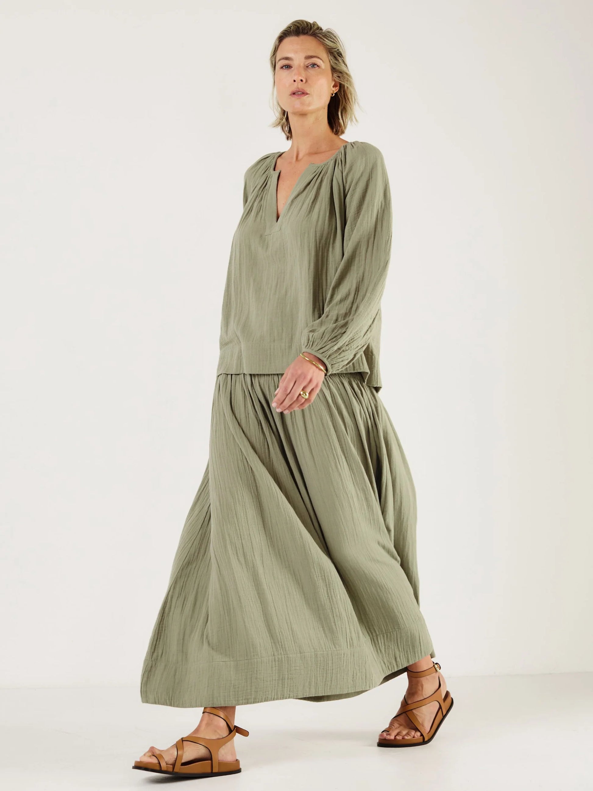 Woman wearing a green outfit, including shirt from Mondo Corsini, on a white background. Side view.