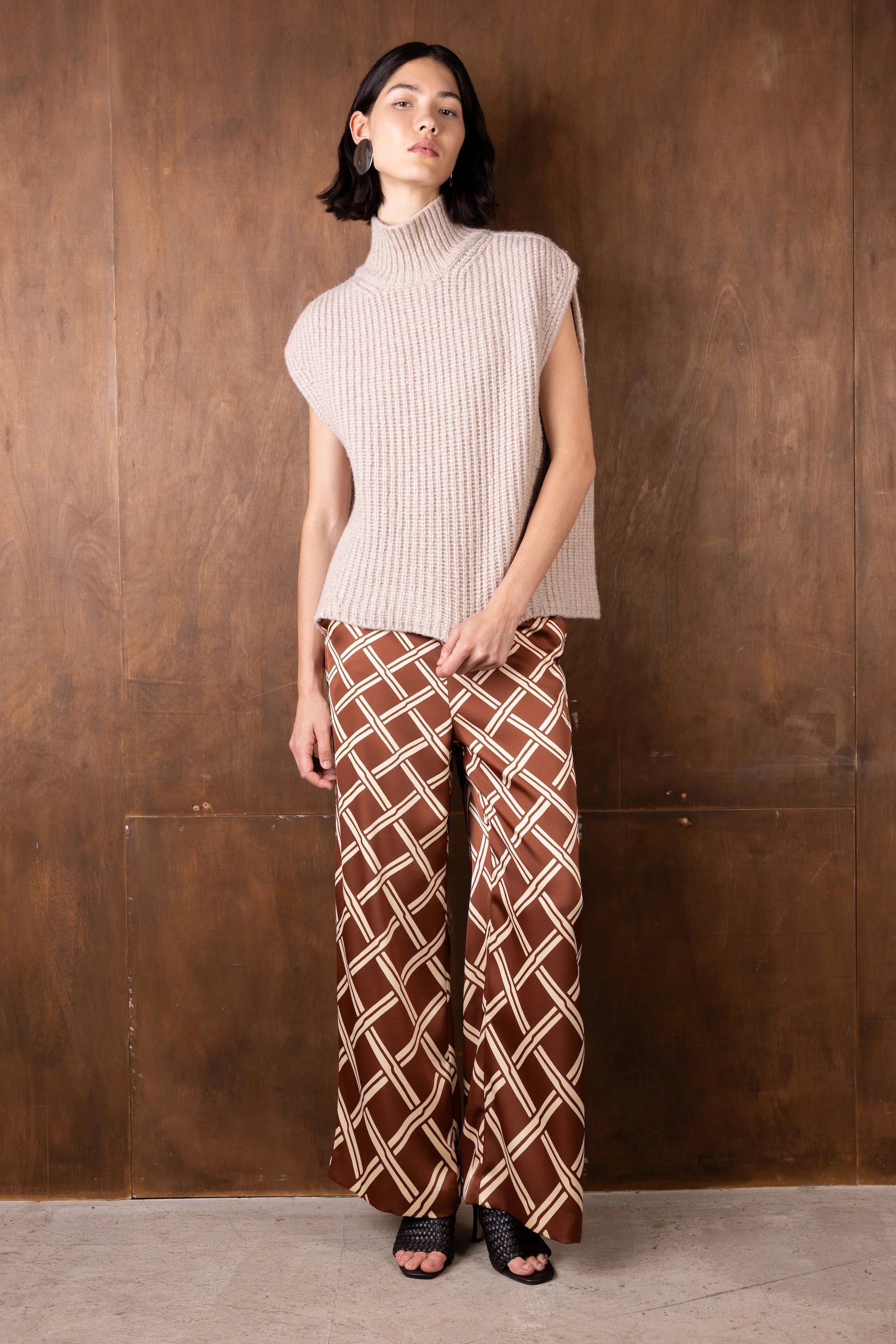 Woman wearing a sleeveless ribbed knit top and patterned pants against a wooden wall.