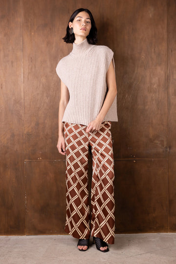 Woman wearing a sleeveless ribbed knit top and patterned pants against a wooden wall.