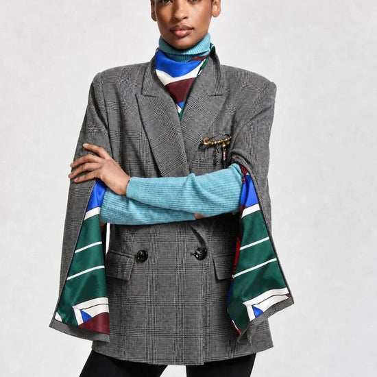 A model wearing a gray double-breasted blazer with a colorful striped scarf draped over the sleeves, styled over a light blue turtleneck sweater and black pants, against a light gray studio background.