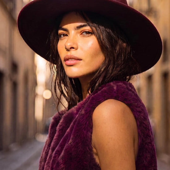 A woman wearing a wide-brimmed burgundy hat and a matching fuzzy sleeveless vest, photographed outdoors on a sunlit city street.