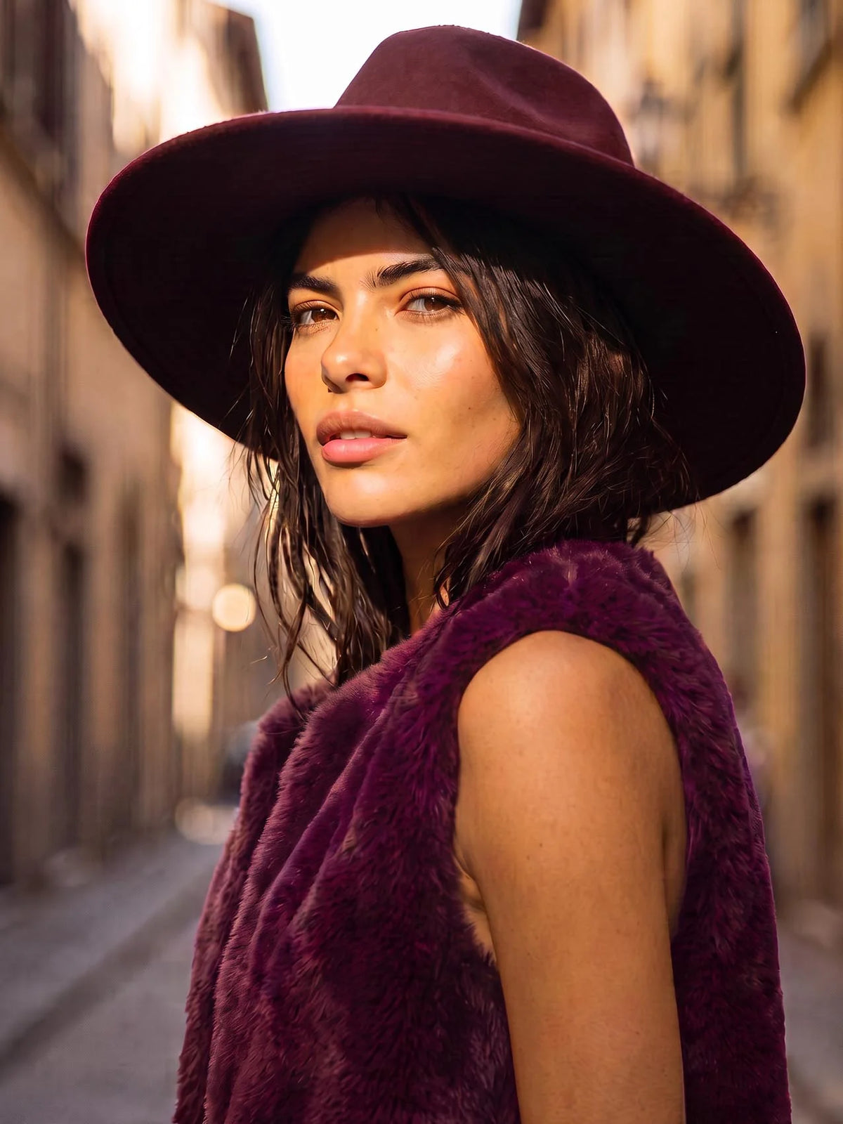 A woman wearing a wide-brimmed burgundy hat and a matching fuzzy sleeveless vest, photographed outdoors on a sunlit city street.