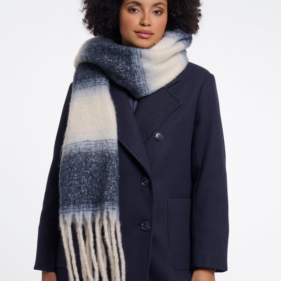 A woman with an afro hairstyle wearing a navy blue coat and a thick knit scarf with a blue and cream ombre gradient pattern and fringe, photographed against a plain white studio background.