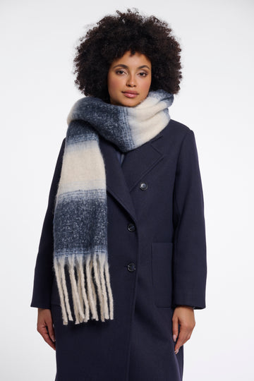 A woman with an afro hairstyle wearing a navy blue coat and a thick knit scarf with a blue and cream ombre gradient pattern and fringe, photographed against a plain white studio background.