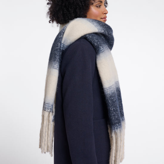 A woman with an afro hairstyle is shown from the side profile wearing a navy blue coat and a thick knit scarf with a blue and cream ombré pattern and fringe ends, set against a plain light gray studio background.
