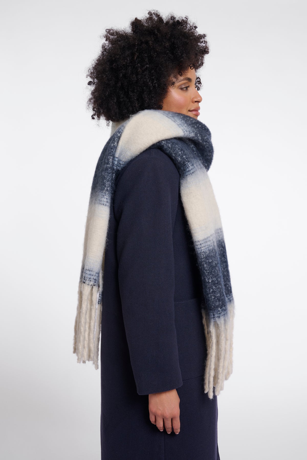 A woman with an afro hairstyle is shown from the side profile wearing a navy blue coat and a thick knit scarf with a blue and cream ombré pattern and fringe ends, set against a plain light gray studio background.