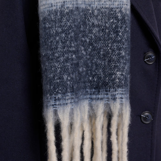 A close-up of a textured knit scarf with a horizontal gradient transitioning from cream at the top to medium blue in the middle and cream fringe at the bottom, worn over a dark navy coat.