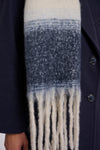 A close-up of a textured knit scarf with a horizontal gradient transitioning from cream at the top to medium blue in the middle and cream fringe at the bottom, worn over a dark navy coat.