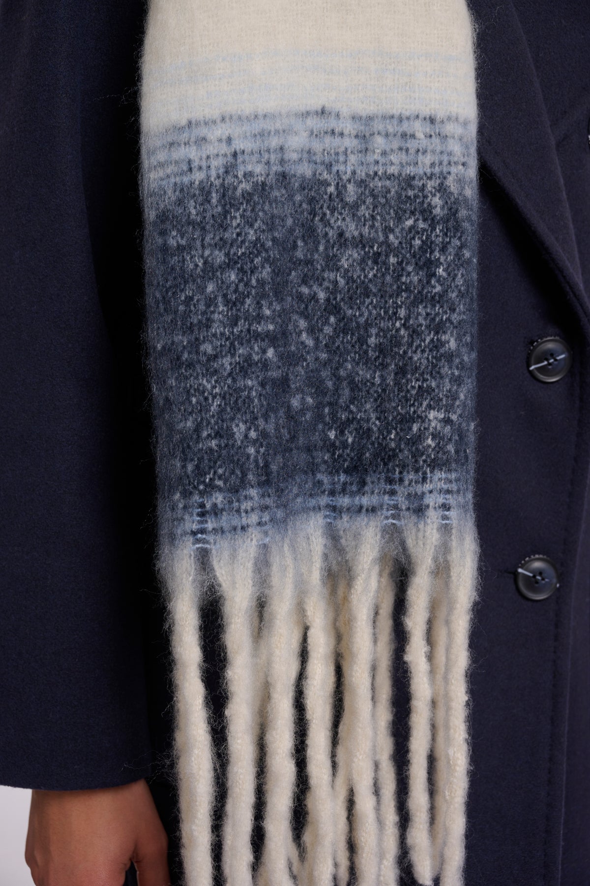 A close-up of a textured knit scarf with a horizontal gradient transitioning from cream at the top to medium blue in the middle and cream fringe at the bottom, worn over a dark navy coat.
