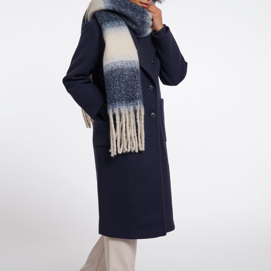 A model wearing a long navy wool coat, a thick blue and cream gradient knit scarf with fringe, and beige wide-leg trousers, standing against a light gray studio background.