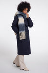 A model wearing a long navy wool coat, a thick blue and cream gradient knit scarf with fringe, and beige wide-leg trousers, standing against a light gray studio background.