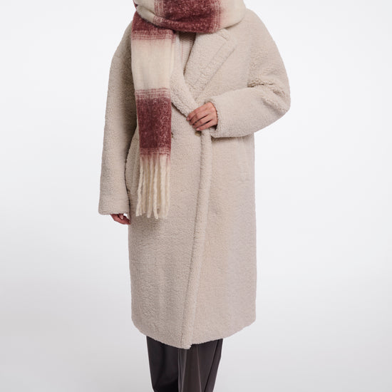 A woman modeling a long cream-colored teddy coat with a chunky knit scarf featuring a cream, rust, and burgundy gradient pattern, set against a plain light gray studio background.