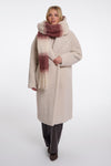 A woman modeling a long cream-colored teddy coat with a chunky knit scarf featuring a cream, rust, and burgundy gradient pattern, set against a plain light gray studio background.