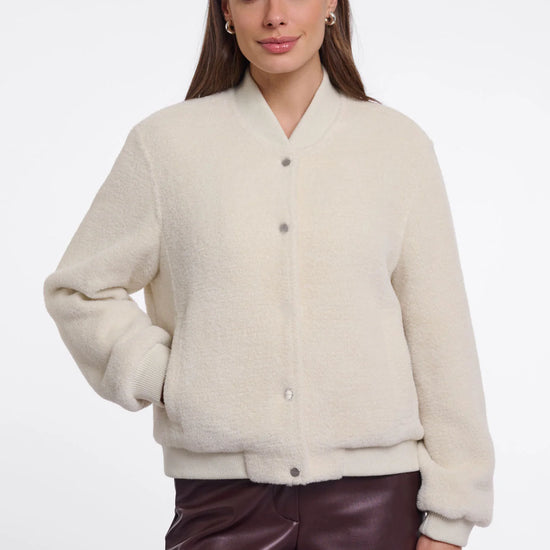 A woman modeling a cream-colored, fuzzy button-up cardigan sweater paired with dark brown faux-leather pants, shot against a plain white studio background.
