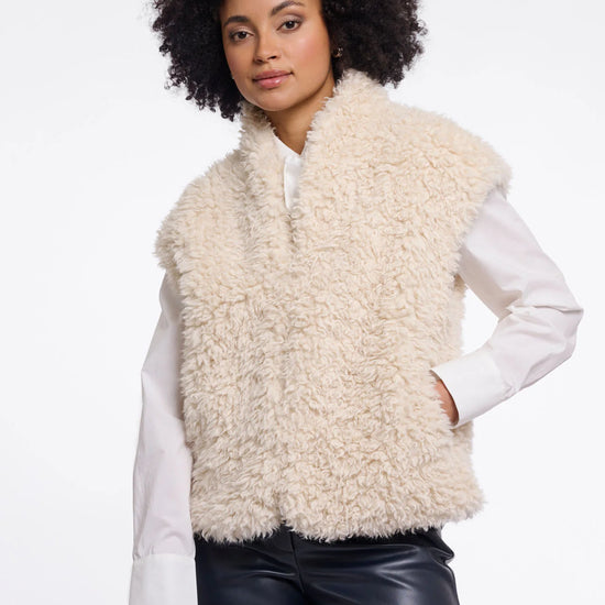 A model with curly black hair wearing a cream-colored curly faux fur vest over a white long-sleeve shirt and black leather pants, photographed against a solid white background.