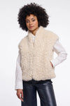 A model with curly black hair wearing a cream-colored curly faux fur vest over a white long-sleeve shirt and black leather pants, photographed against a solid white background.