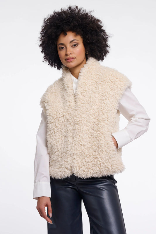 A model with curly black hair wearing a cream-colored curly faux fur vest over a white long-sleeve shirt and black leather pants, photographed against a solid white background.