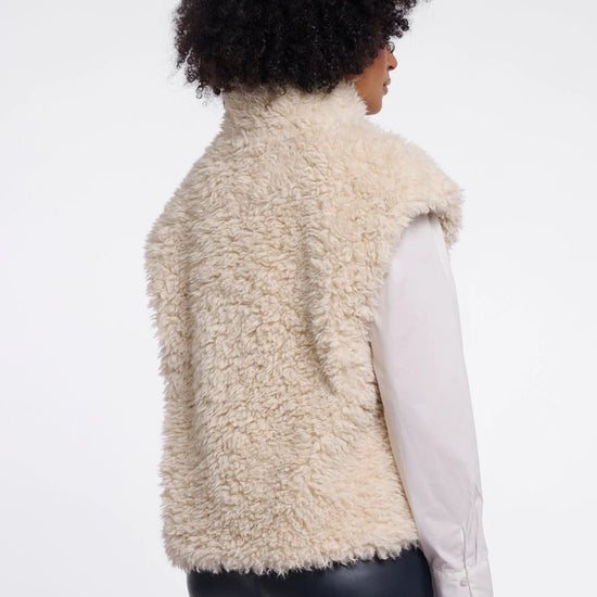 A person shown from the back wearing a cream-colored curly shearling-style sleeveless vest over a white long-sleeve shirt and dark pants, against a plain light gray studio background.