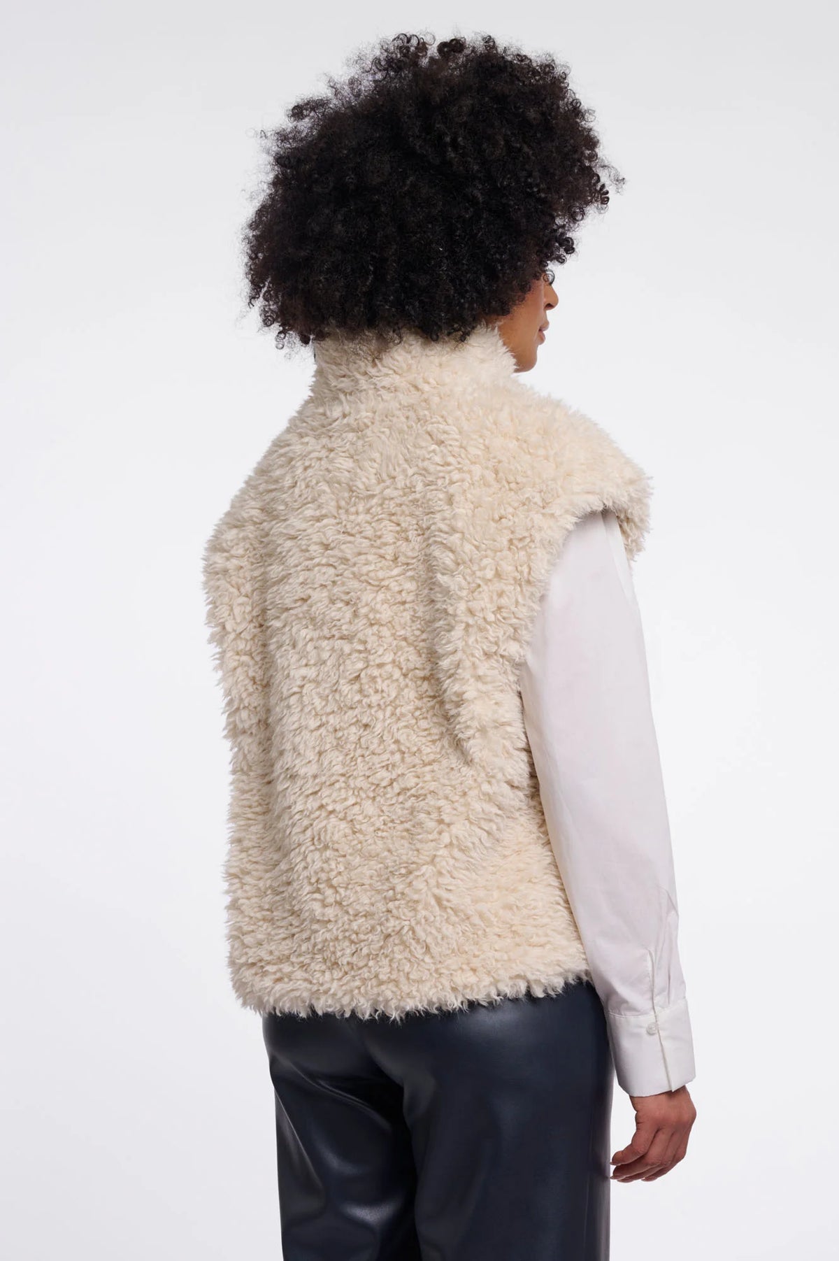 A person shown from the back wearing a cream-colored curly shearling-style sleeveless vest over a white long-sleeve shirt and dark pants, against a plain light gray studio background.
