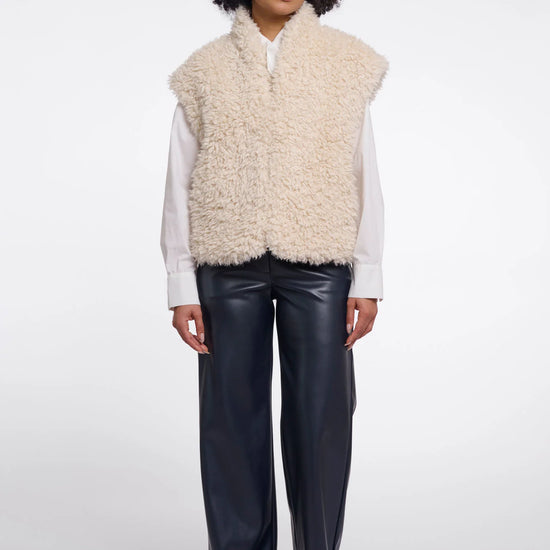 A model wearing a cream-colored curly faux fur vest over a white long-sleeve shirt and black wide-leg leather pants, standing against a light gray studio background.