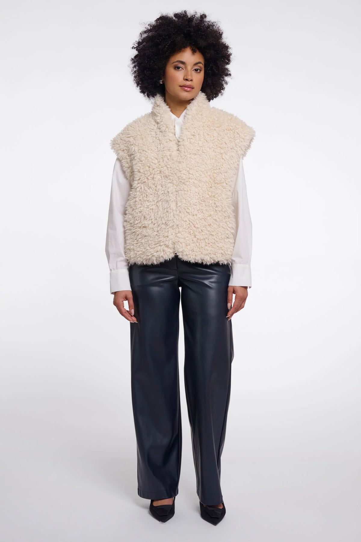 A model wearing a cream-colored curly faux fur vest over a white long-sleeve shirt and black wide-leg leather pants, standing against a light gray studio background.
