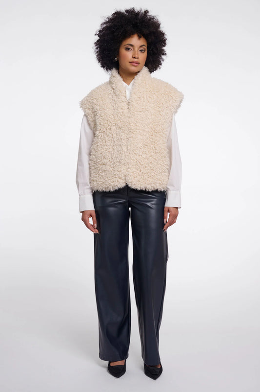A model wearing a cream-colored curly faux fur vest over a white long-sleeve shirt and black wide-leg leather pants, standing against a light gray studio background.