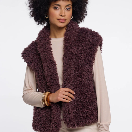 A woman with a large black afro wearing a shaggy, sleeveless plum-brown vest over a cream long-sleeve top and cream pants, photographed against a light gray studio background.