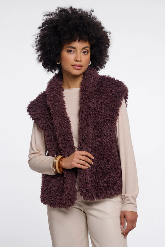 A woman with a large black afro wearing a shaggy, sleeveless plum-brown vest over a cream long-sleeve top and cream pants, photographed against a light gray studio background.