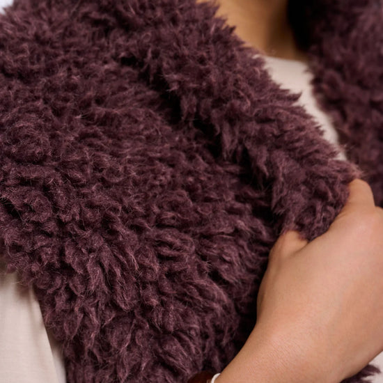 A close-up of a person wearing a chunky, shaggy faux-fur vest in a deep plum purple color, paired with a cream-colored top and wooden bangles.
