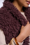 A close-up of a person wearing a chunky, shaggy faux-fur vest in a deep plum purple color, paired with a cream-colored top and wooden bangles.