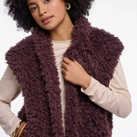 A woman with an afro hairstyle wearing a chunky, shaggy, deep plum-brown faux fur vest over a light beige long-sleeve top, photographed against a solid white studio background.