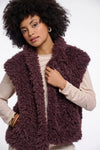 A woman with an afro hairstyle wearing a chunky, shaggy, deep plum-brown faux fur vest over a light beige long-sleeve top, photographed against a solid white studio background.