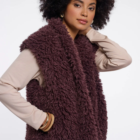 A woman with an afro hairstyle wearing a shaggy, textured plum-brown faux fur vest over a beige long-sleeve top and beige pants, posed against a solid white studio background.