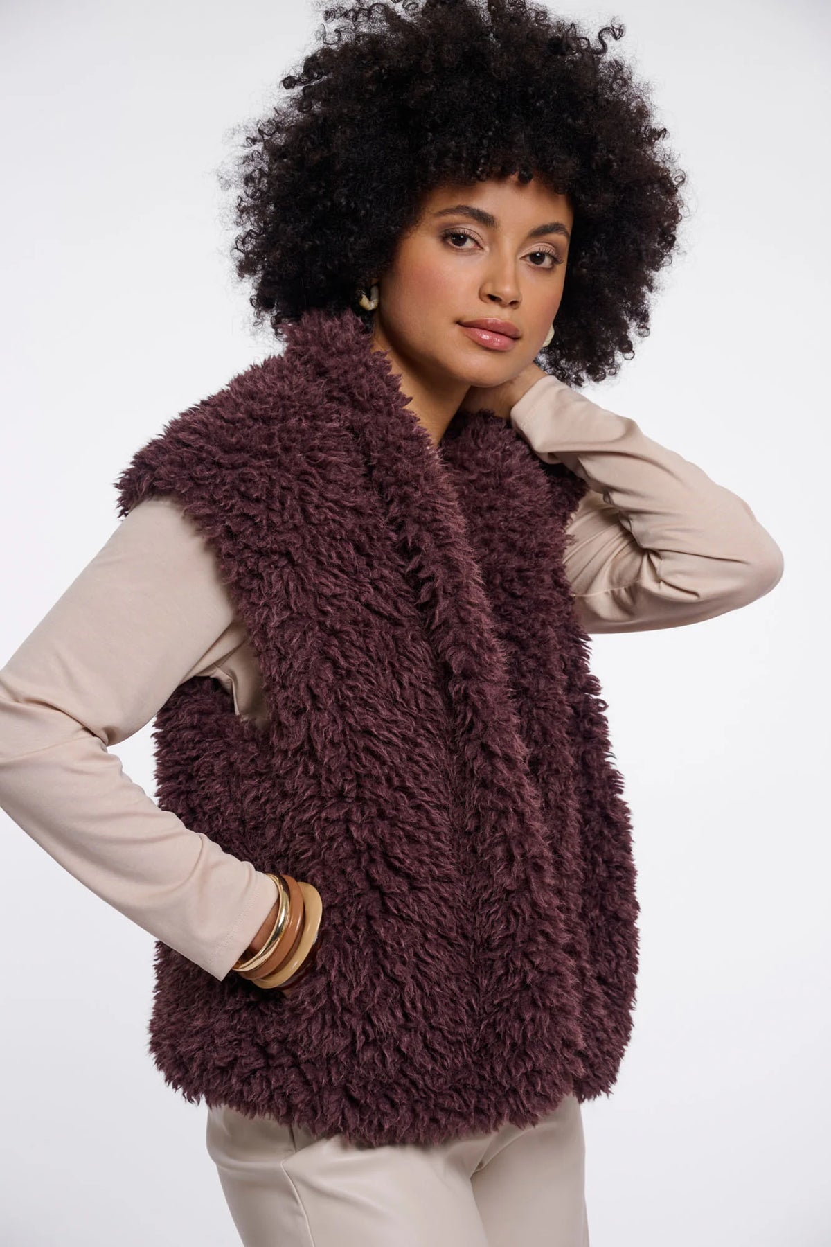 A woman with an afro hairstyle wearing a shaggy, textured plum-brown faux fur vest over a beige long-sleeve top and beige pants, posed against a solid white studio background.