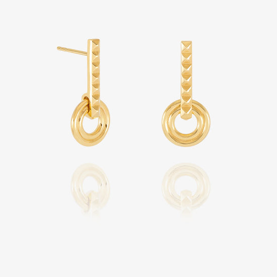 A pair of gold-toned drop earrings with a textured vertical bar, a circular ring, and a smaller concentric ring, displayed on a clean white background with a soft reflection below.