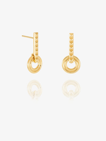 A pair of gold-toned drop earrings with a textured vertical bar, a circular ring, and a smaller concentric ring, displayed on a clean white background with a soft reflection below.