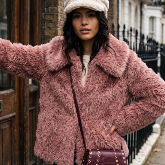 A woman wearing a dusty pink faux fur jacket, a cream-colored newsboy cap, and a dark burgundy studded crossbody bag, posing on a city street.