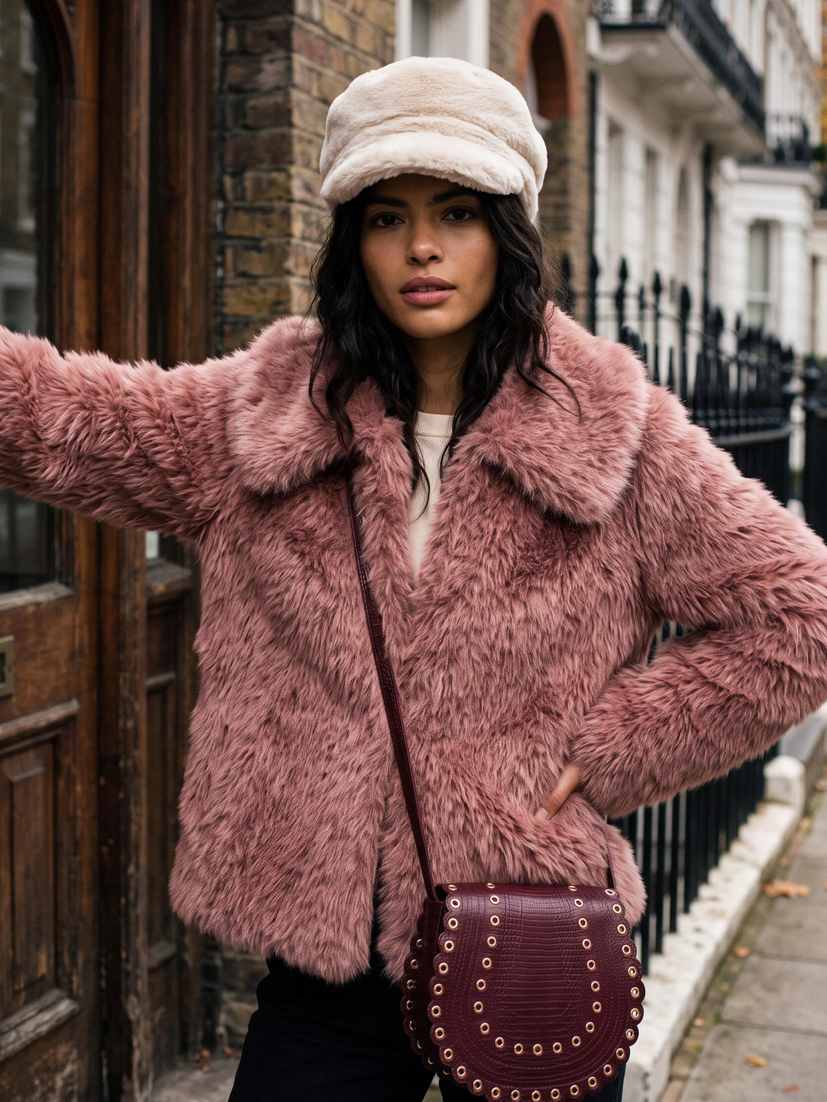 A woman wearing a dusty pink faux fur jacket, a cream-colored newsboy cap, and a dark burgundy studded crossbody bag, posing on a city street.